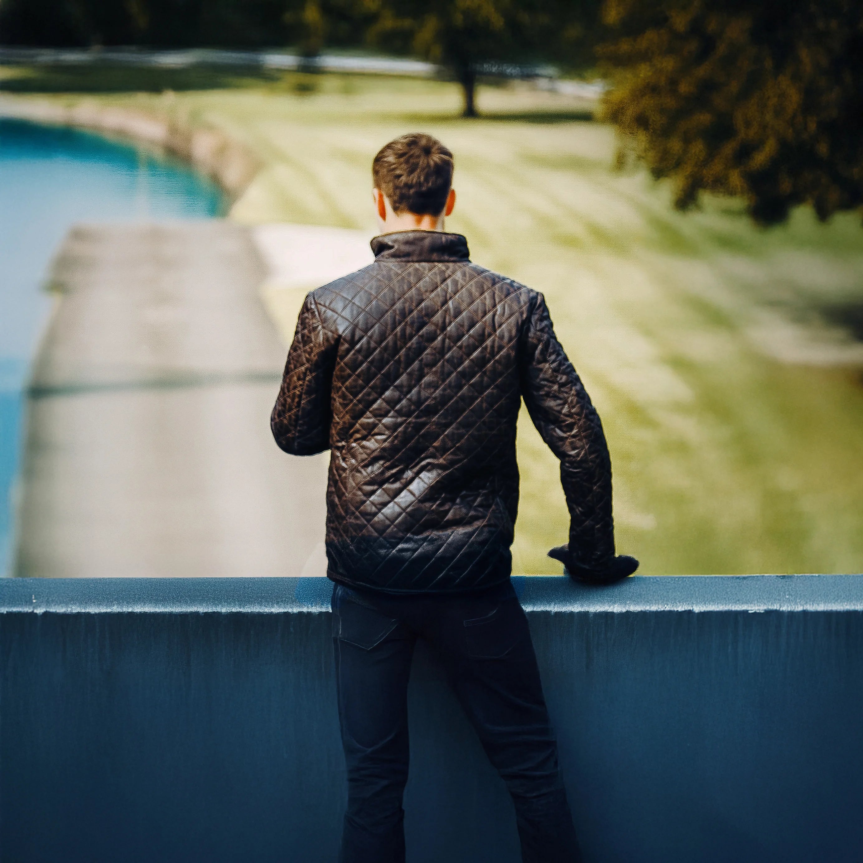 Man in a quilted jacket standing on a bridge over a body of water with trees in the background